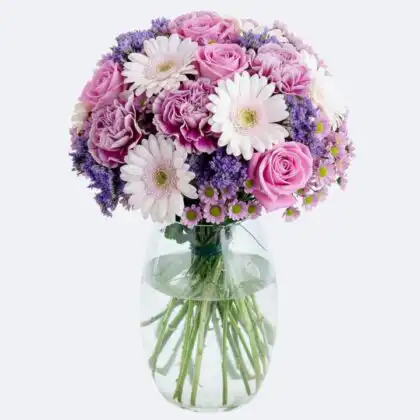 A vibrant gerbera rose bouquet with pink roses, white gerberas, and purple carnations arranged in a clear glass vase.