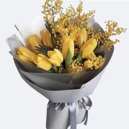 A stunning Yellow Tulips Bouquet accented with golden mimosa and elegant silver gift wrapping.