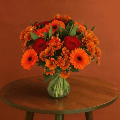 Autumn Flower Vase Arrangement featuring vibrant orange gerbera daisies, rich red roses, and seasonal chrysanthemums on a wooden table.