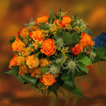 A vibrant bouquet of deep orange Autumn Roses Hand Tied with thistle and yellow accents, set against a blurred background of fall colors.