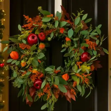 A close-up shot of a rustic, handmade autumn door wreath featuring faux orange, red, and yellow leaves and small berries.