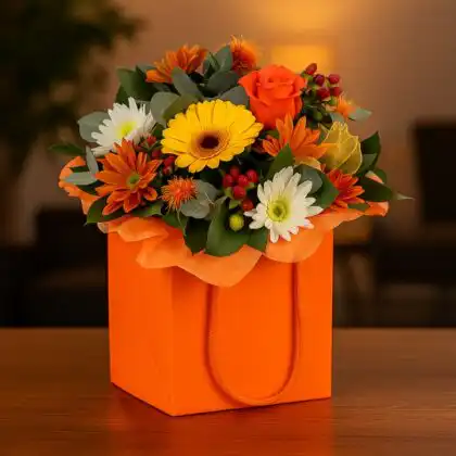 A brightly colored orange gift bag containing a beautiful Handmade Autumn Flower Box arrangement of yellow gerbera, orange roses, and fall foliage.