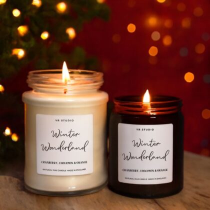 A set of two burning luxury Christmas scented candles in glass jars, scented with Cranberry, Cinnamon & Orange, set against holiday lights.