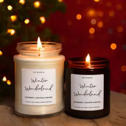 A set of two burning luxury Christmas scented candles in glass jars, scented with Cranberry, Cinnamon & Orange, set against holiday lights.