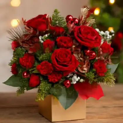 Festive flowers for Christmas delivery featuring red roses, carnations, red baubles, and pine sprigs in a golden decorative box, with a blurry Christmas tree background. Luxury Christmas Hat Box Flowers for sale