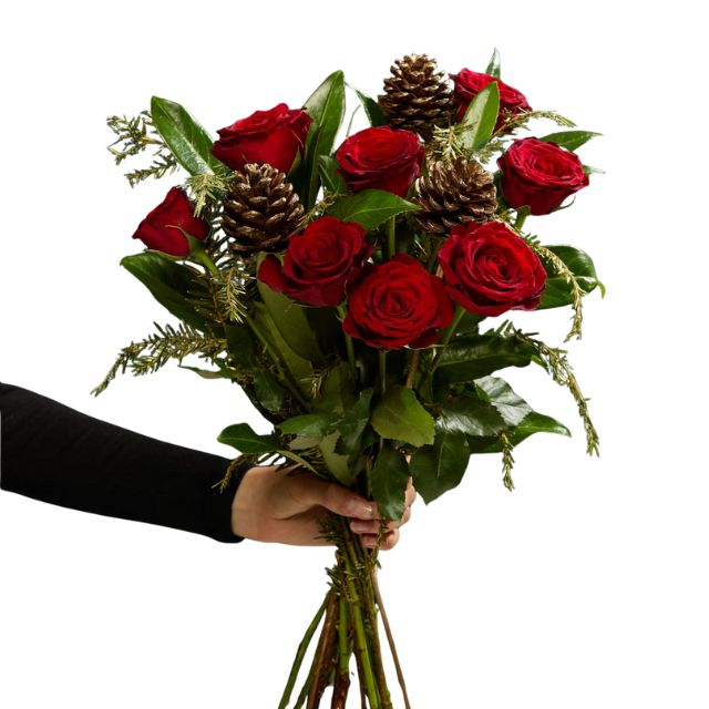 Blog 10 A hand holding a vibrant bouquet of deep red roses mixed with pine sprigs and natural pine cones, perfect for holiday flower arrangements Christmas. Red Rose Christmas Bouquet for sale