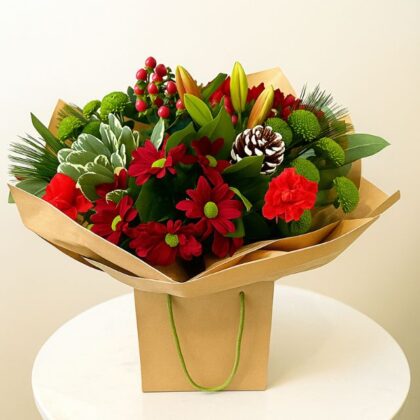 Festive bouquet featuring red and green chrysanthemums, lilies, red berries, pine needles, and a small pine cone, presented in a brown paper gift bag. Unique Christmas flower arrangements
