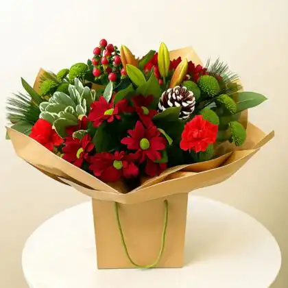 Festive bouquet featuring red and green chrysanthemums, lilies, red berries, pine needles, and a small pine cone, presented in a brown paper gift bag. Unique Christmas flower arrangements