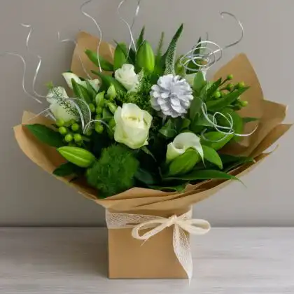 Hand-tied Christmas rose and lily bouquet with white flowers, green foliage, silver accents, and a pinecone in a brown kraft gift box. Christmas bouquet delivery
