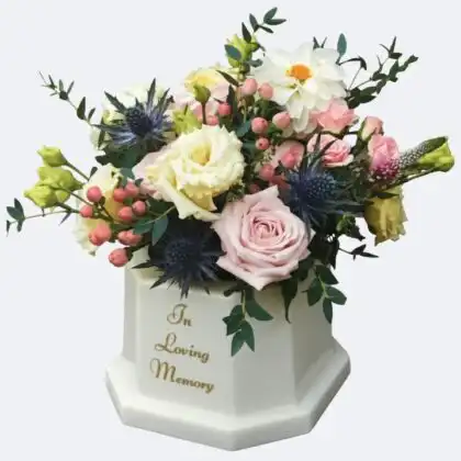 In Loving Memory Flower Vase featuring a graceful assortment of blush pink roses, creamy yellow blooms, striking blue thistles, and complementary greenery, displayed in a white keepsake memorial urn.