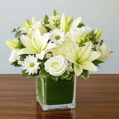 White sympathy flowers with lilies and roses in a glass cube lined with green leaves for funeral tribute.
