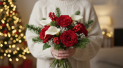 A person in a white sweater holds a festive bouquet of red roses, white calla lilies, and red berries, a popular example of What Flower Is Given the Most at Christmas.